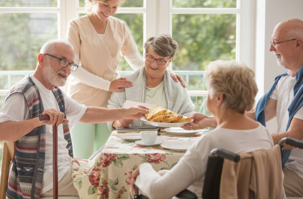 老人ホームダイニングルームでテーブルに座って、ケーキを食べて、役に立つ介護者の先輩友達のグループ - senior adult nursing home eating home interior ストックフォトと画像