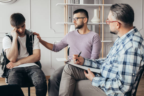 young man in glasses comforting his depressed friend during meeting with counselor - mental health professional family couple psychiatrist imagens e fotografias de stock