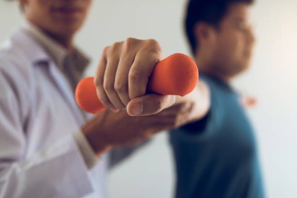 paciente de ajuda masculino novo asiático do fisioterapeuta com exercícios de levantamento dos dumbbells no escritório. - pain the human body physical therapy human joint - fotografias e filmes do acervo