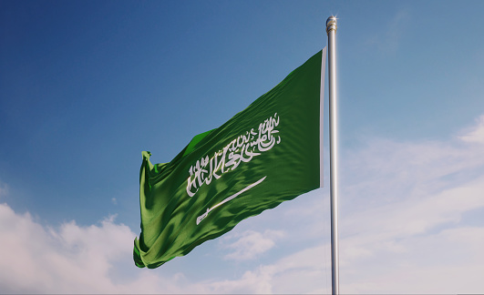 The Saudi national flag flutters in the wind against a clear blue sky with sunlight