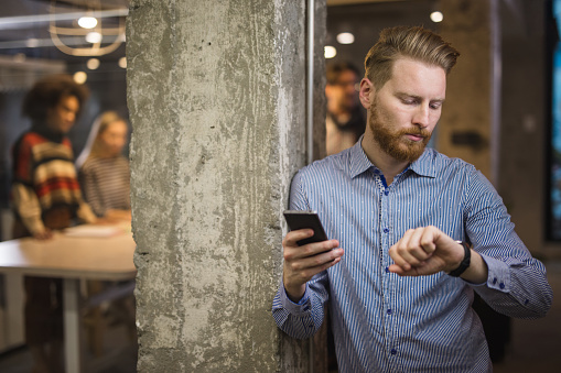 Redhead businessman looking at time on his wristwatch while using mobile phone in the office. There are people in the background.