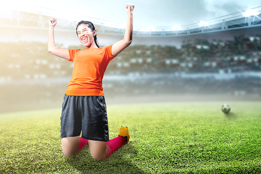 Asian football player woman celebrate her goal with raised arms and kneeling on the football field at stadium