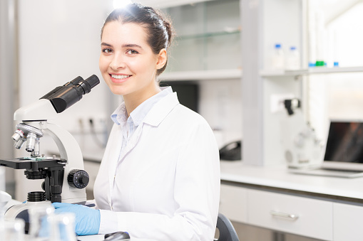 Portrait of smiling attractive female university student of biological department sitting at desk and using microscope in scientific laboratory