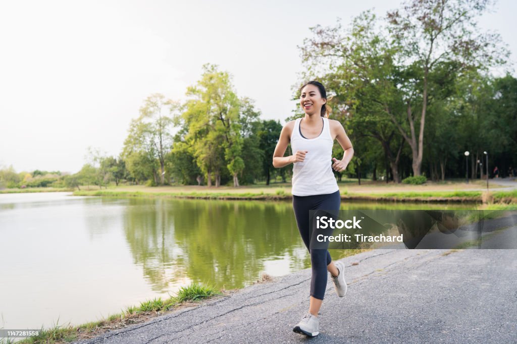 Gesunde schöne junge asiatische Läuferin in Sportkleidung Laufen und Joggen auf dem Bürgersteig in der Nähe des Sees am Park am Morgen. Lifestyle-Fitness und aktive Frauen trainieren im Stadtkonzept. - Lizenzfrei Rennen - Körperliche Aktivität Stock-Foto