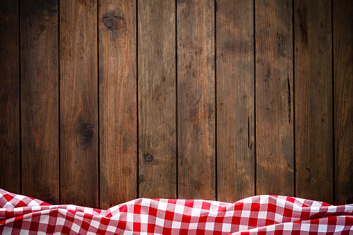 Backgrounds: high angle view of an empty brown wooden table with vertical stripes. A crumpled red and white gingham tablecloth is at the edge of the bottom border of an horizontal frame leaving a useful copy space for text, logo or product montage. Predominant colors are brown and red. Low key DSRL indoors photo taken with Canon EOS 5D Mk II and Canon EF 24-105mm f/4L IS USM Wide Angle Zoom Lens