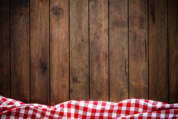 Empty brown wooden table with gingham tablecloth at the top border. Copy space available. Backgrounds: high angle view of an empty brown wooden table with vertical stripes. A crumpled red and white gingham tablecloth is at the edge of the bottom border of an horizontal frame leaving a useful copy space for text, logo or product montage. Predominant colors are brown and red. Low key DSRL indoors photo taken with Canon EOS 5D Mk II and Canon EF 24-105mm f/4L IS USM Wide Angle Zoom Lens high section stock pictures, royalty-free photos & images