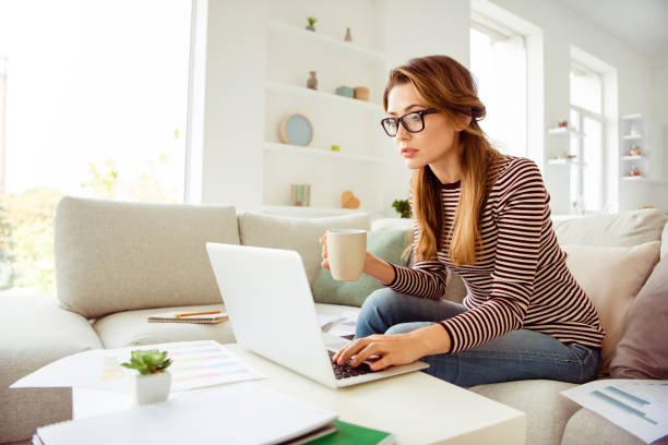 istock Porrait of minded female beautiful worker have graphs report solve decide choose solution use user device take mug beverage wear specs striped shirt long wavy curly hair sit divan in living room 1147283292