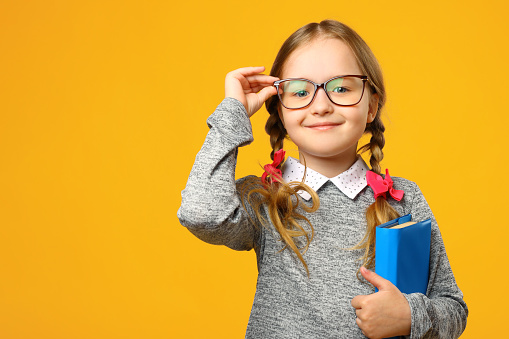 Portrait of a cute little kid girl on a yellow background. Child schoolgirl looking at the camera, holding a book and straightens glasses. The concept of education. Copy space.