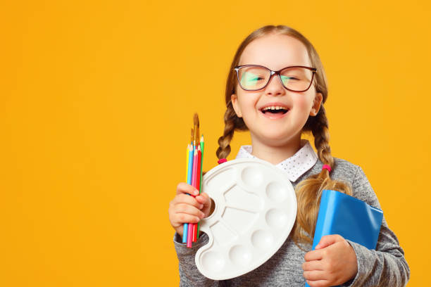 Portrait of a cheerful little child girl on a yellow background. Schoolgirl holds a book, pencils, brushes and a palette. The concept of education. Portrait of a cheerful little child girl on a yellow background. Schoolgirl holds a book, pencils, brushes and a palette. The concept of education. child paintbrush stock pictures, royalty-free photos & images