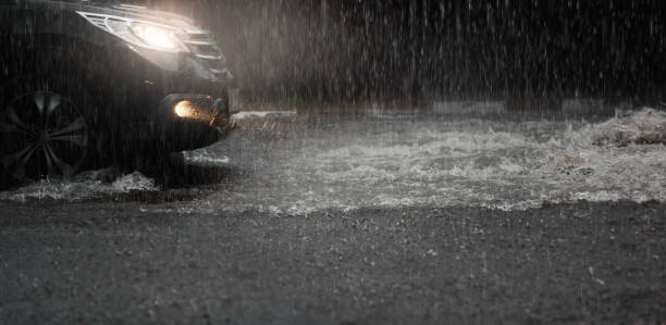 o carro com faróis corre através da água da inundação após a queda dura da chuva na noite. - luz de veículo - fotografias e filmes do acervo