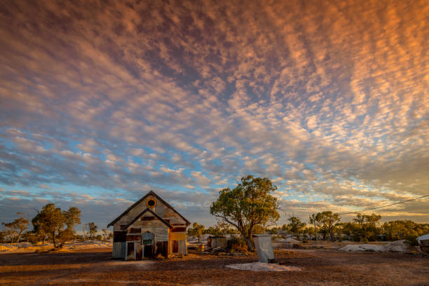 Rusty old corrugated iron church below mackerel sky of orange glowing Stratocumulus clouds at dusk. The church, once part of a film set, sits deserted on the dry opal fields at Lightning Ridge, a small town in northern New South Wales, Australia. A small corrugated metal dunny (toilet) stands nearby.