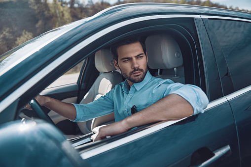 Handsome young man in smart casual wear looking away while driving a status car