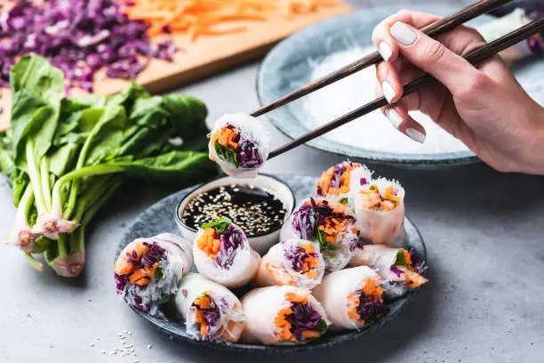 Eating Asian Cuisine Spring Rolls Or Rice Paper Rolls With Vegetables And Shrimps. Hand Holding Roll With Chopsticks