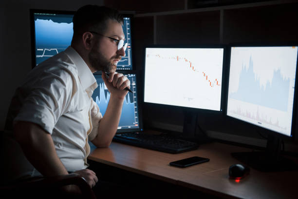 istock Thoughtful look. Bearded man in white shirt works in the office with multiple computer screens in index charts 1147314627