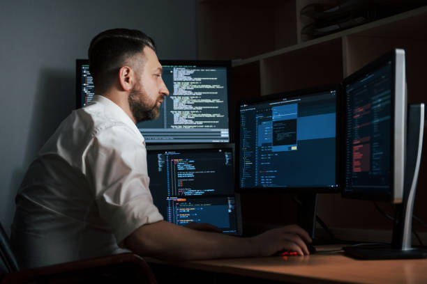 istock Adult experienced programmer. Bearded man in white shirt works in the office with multiple computer screens in index charts 1147315261