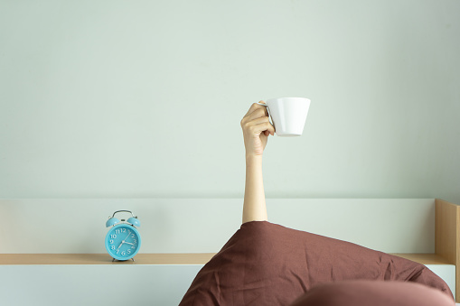 Woman showing arm raised up holding coffee cup on bed under duvet in the bed room, Young girl with hand sticking out from the blanket holding tea cup. wake up drinking coffee in morning concept.