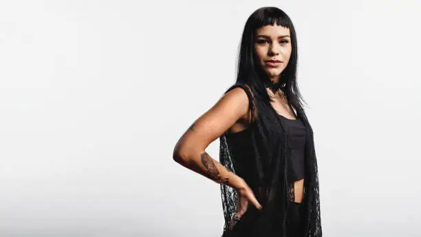 Woman with fashion piercing on lips standing against white background with hand on waist. Portrait of a young gothic woman with tattoo on hand looking at camera.