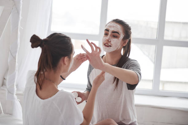 istock Sitting against white window. Conception of skin care by using white mask on the face. Two female sisters have weekend at bedroom 1147328305