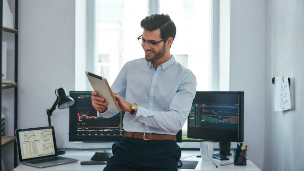 Good news. Happy young trader in formal wear is looking is using his digital tablet and smiling while standing in front of computer screens with trading charts in the office. stock photo