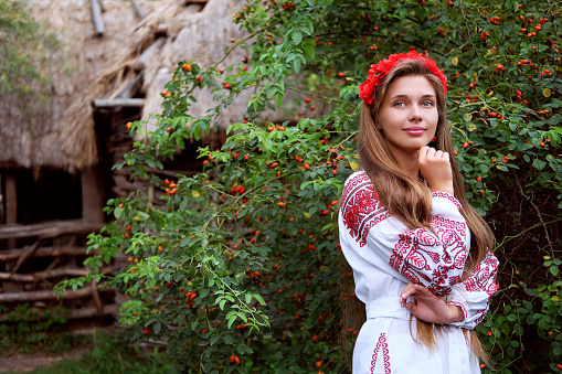 Beautiful slavonic girl with long blond hair and blue eyes with flower crown in white and red embroidered costume at the gate looking welcome.Traditional clothes of Ukrainian region.