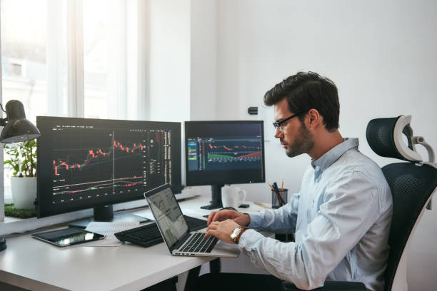 istock Workplace of trader. Young bearded trader wearing eyeglasses using his laptop while sitting in office in front of computer screens with trading charts and financial data 1147352112