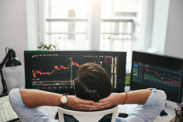 istock Time to relax. Back view of young businessman or trader holding hands behind head while looking at charts and financial data on computers in his modern office. 1147352129