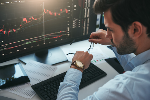 Time to trade. Bearded male trader looking at watch on his hand while working with data and charts on computer screens in his modern office. Stock exchange. Trade concept. Investment concept
