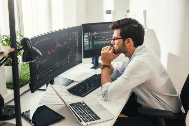 Successful trader. Back view of bearded stock market broker in eyeglasses analyzing data and graphs on multiple computer screens while sitting in modern office. stock photo