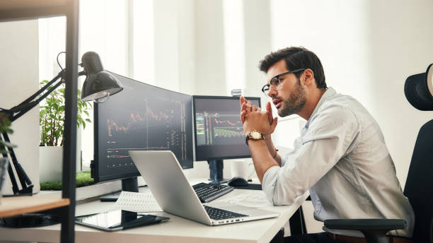 istock Trading strategy. Successful young trader in eyeglasses looking at analyzing trading charts on computer screens in while sitting in his modern office. 1147352185