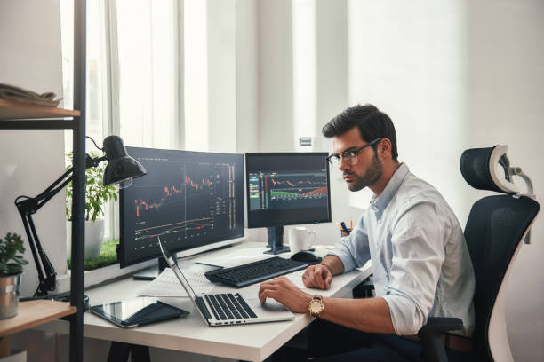 istock Busy working day. Young bearded trader in eyeglasses working with laptop while sitting in his modern office in front of computer screens with trading charts. 1147352265