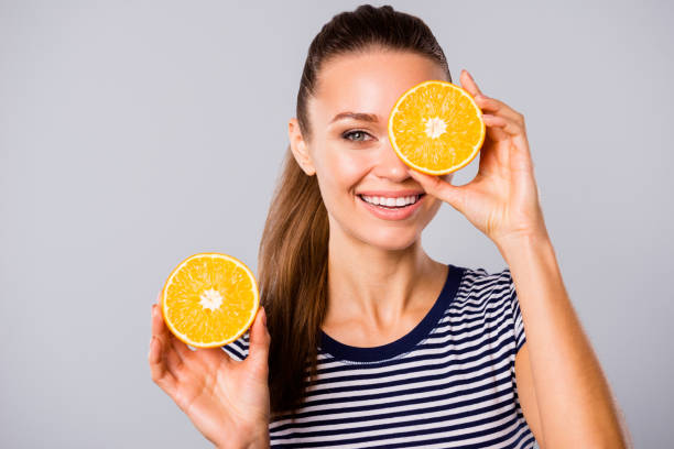 Close up photo beautiful amazing she her lady hold hands arms big large two half slices orange hide one eye new oil extract wash lotion wear blue white striped t-shirt isolated grey background Close up photo beautiful amazing she her lady hold hands arms big large two half slices orange hide one eye new oil extract wash lotion wear blue white striped t-shirt isolated grey background. oranges with faces stock pictures, royalty-free photos & images