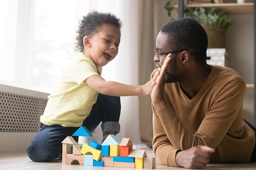 African American father in glasses with toddler son playing with colorful wooden constructor, giving five, adorable little child and black dad or babysitter having fun on warm floor at home