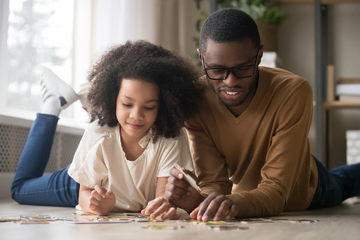 Happy African American father in glasses with preschool daughter assembling jigsaw puzzle, smiling dad and adorable girl child playing on warm floor at home together, family weekend, close up