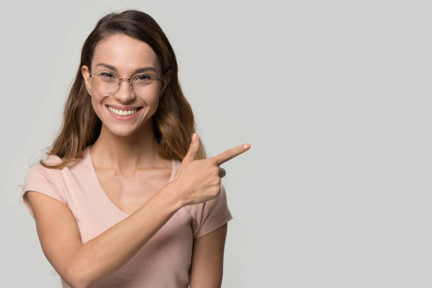 Friendly woman in glasses pointing finger to copyspace studio shot On grey wall background positive smiling female in glasses looking at camera pointing finger aside at copy space, client recommending eyewear great offer store, freespace for your advertisement text beautiful women giving head stock pictures, royalty-free photos & images