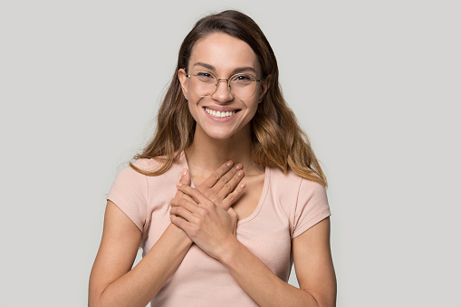 Head shot studio portrait of pretty woman in glasses looking at camera smiling feels grateful for love care and support holding hands on chest sincere feelings from heart concept, over grey background