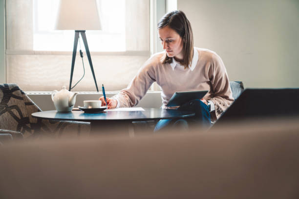 A girl writing down information she had found on her tablet A young student with short brown hair and a pink jumper sitting in a coffeeshop, using her tablet to find information and writing in down on a piece of paper. education student mobile phone university stock pictures, royalty-free photos & images