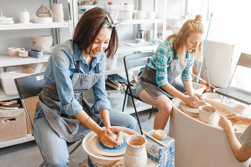 Two girls friends smiling and talking while working on potters wheel making clay handmade craft in pottery workshop, friendship and guidance concept