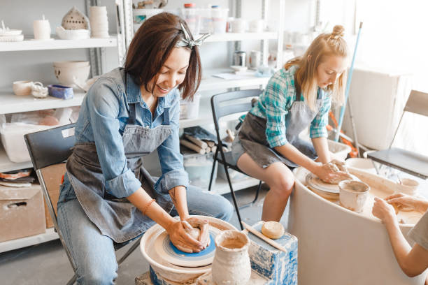 dois amigos das meninas que sorriem e que falam ao trabalhar na roda dos potters que faz a argila handmade ofício na oficina da cerâmica, na amizade e no conceito da orientação - potter human hand craftsperson molding - fotografias e filmes do acervo