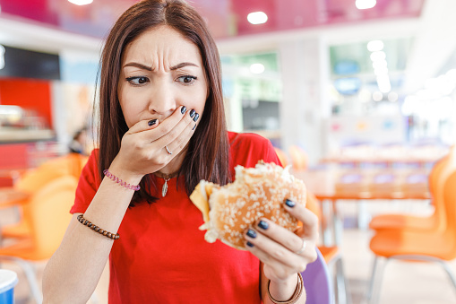 Woman with burger in hand, making bad and disgusting face, concept of the bad spoiled food and problems with digestion