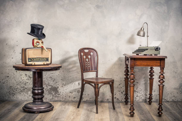 vintage loft room with antique chair, broadcast radio, carnival mask, cylinder hat, old typewriter and lamp on oak wooden desk front concrete wall background with shadows. retro style filtered photo - writing typewriter 1950s style retro revival imagens e fotografias de stock