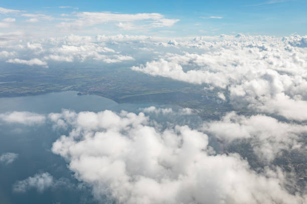 Bird's eye view with the clouds above is captured from an airplane window on a sunny autumn day. The sky on the horizon is blue, and there are a body of water and land below.