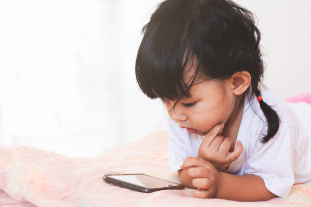 niedliches asiatisches mädchen, das smartphone auf ihrem bett in ihrem zimmer liegt - baby pointing child showing stock-fotos und bilder