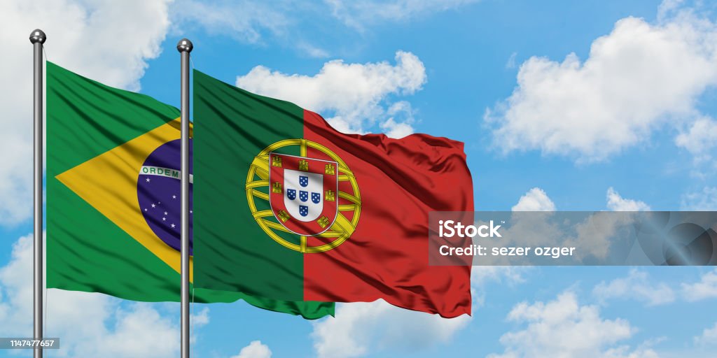 Bandeira de Brasil e de Portugal que acena no vento de encontro ao céu azul nebuloso branco junto. Conceito da diplomacia, relações internacionais. - Foto de stock de Portugal royalty-free