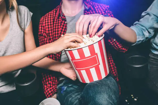 Close up of red with white basket of popcorn that both girl and guy are holding. All of them are taking popcorn out of basket. Ð¡ut view