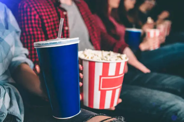 Photo of Close up of tasty but unhealthy food. There are basket of popcorn and a blue cup of coke on picture. Man and woman are holding it together.