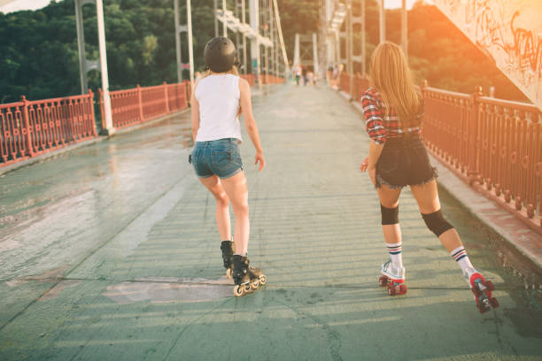 two slim and sexy young women and roller skates. one female has an inline skates and the other has a quad skates. girls ride in the rays of the sun. - roller skate imagens e fotografias de stock