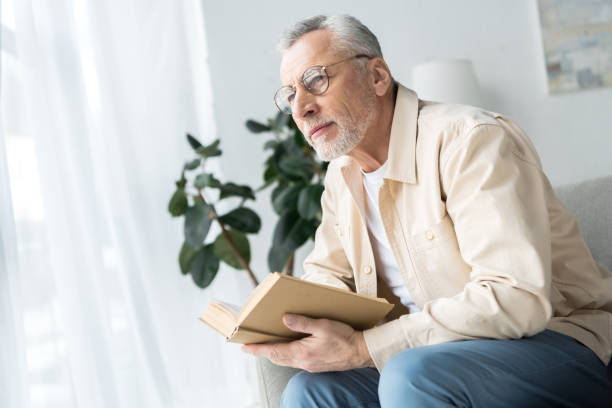 istock senior man in glasses holding book while sitting at home 1147492542
