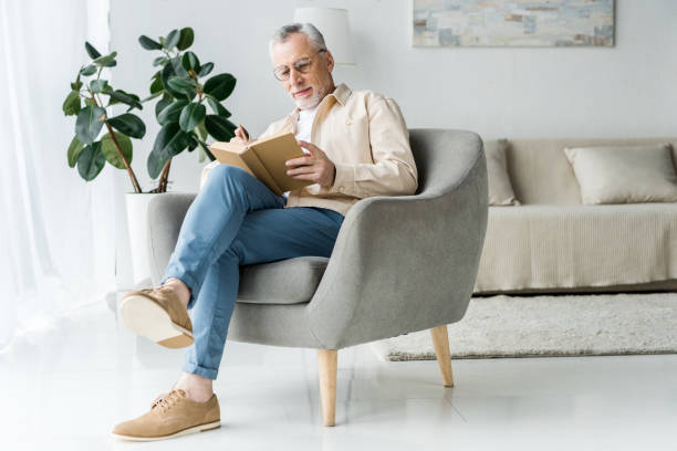 homme aîné dans le livre de lecture de lunettes tandis que assis dans le fauteuil à la maison - vieux fauteuil photos et images de collection