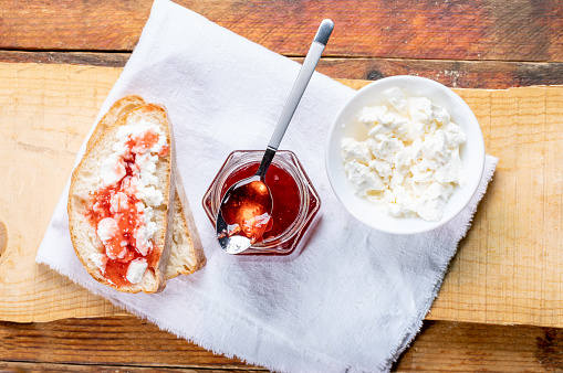 Toasts with curd and strawberry jam, open glass jar full of homemade strawberry  jam with spoon and white bowl with curd on white napkin on wooden plank on old rustic wooden table. Natural homemade breakfast table. Top view, flat lay.