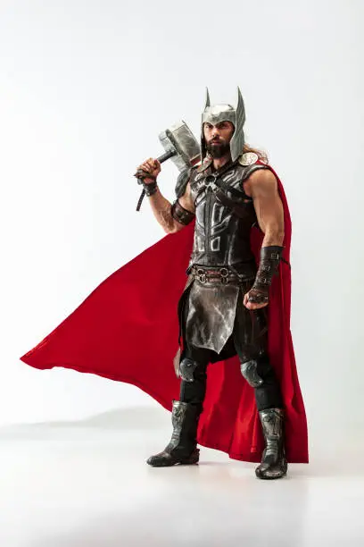 Long hair and muscular male model in leather viking's costume with the big hammer cosplaying Thor isolated on white studio background. Full-lenght portrait. Fantasy warrior, antique battle concept.